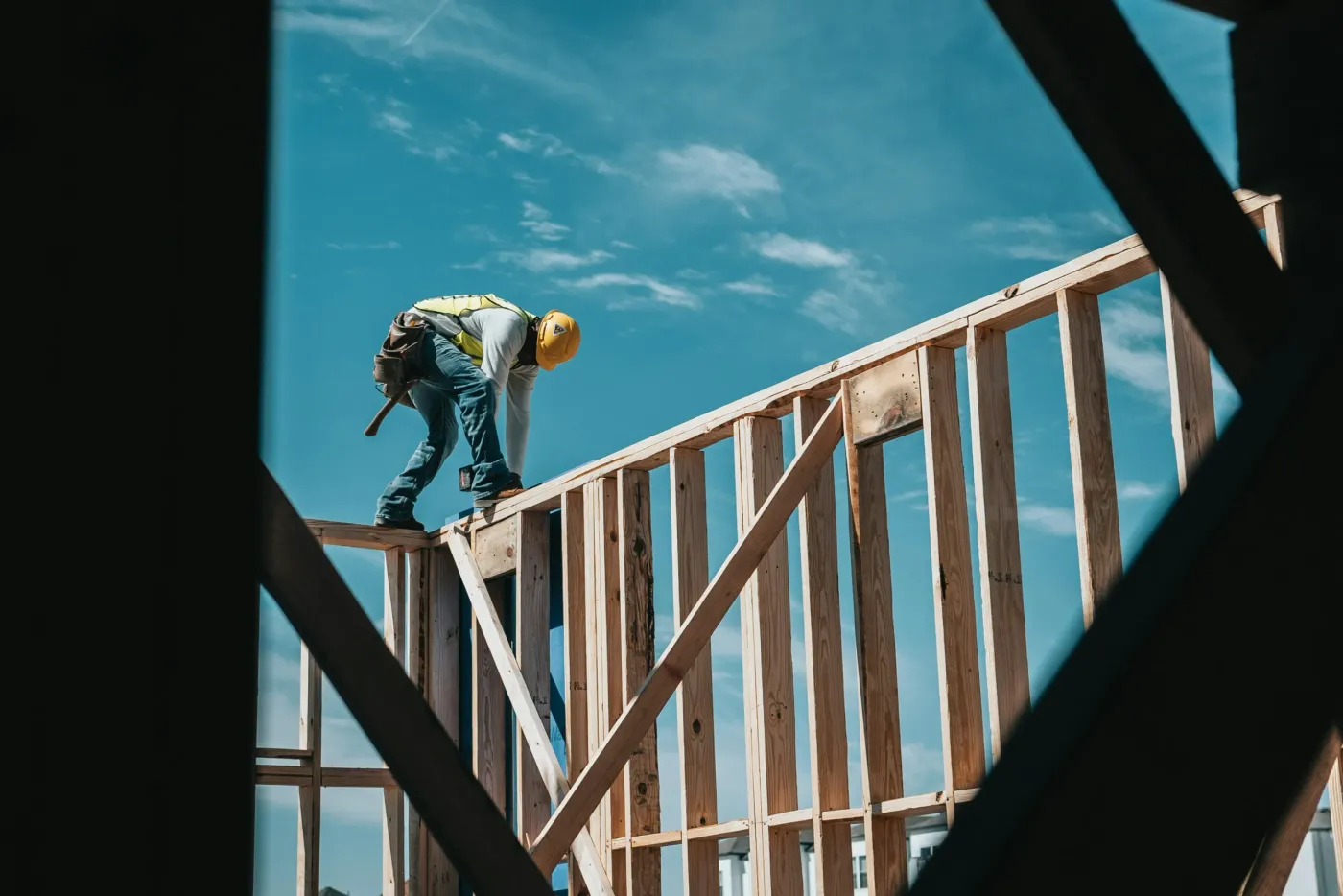 Builder in a hard hat and hi-vis vest standing on top of a new timber stud wall frame against a blue sky