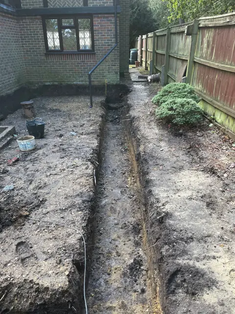 A foundation trench dug along the side of the house, beside the garden fence