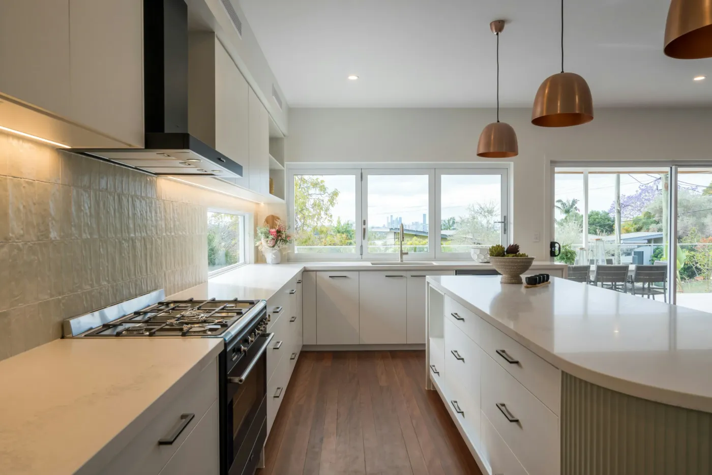 White handleless kitchen with a curved island, copper pendant lights and folding windows over the sink