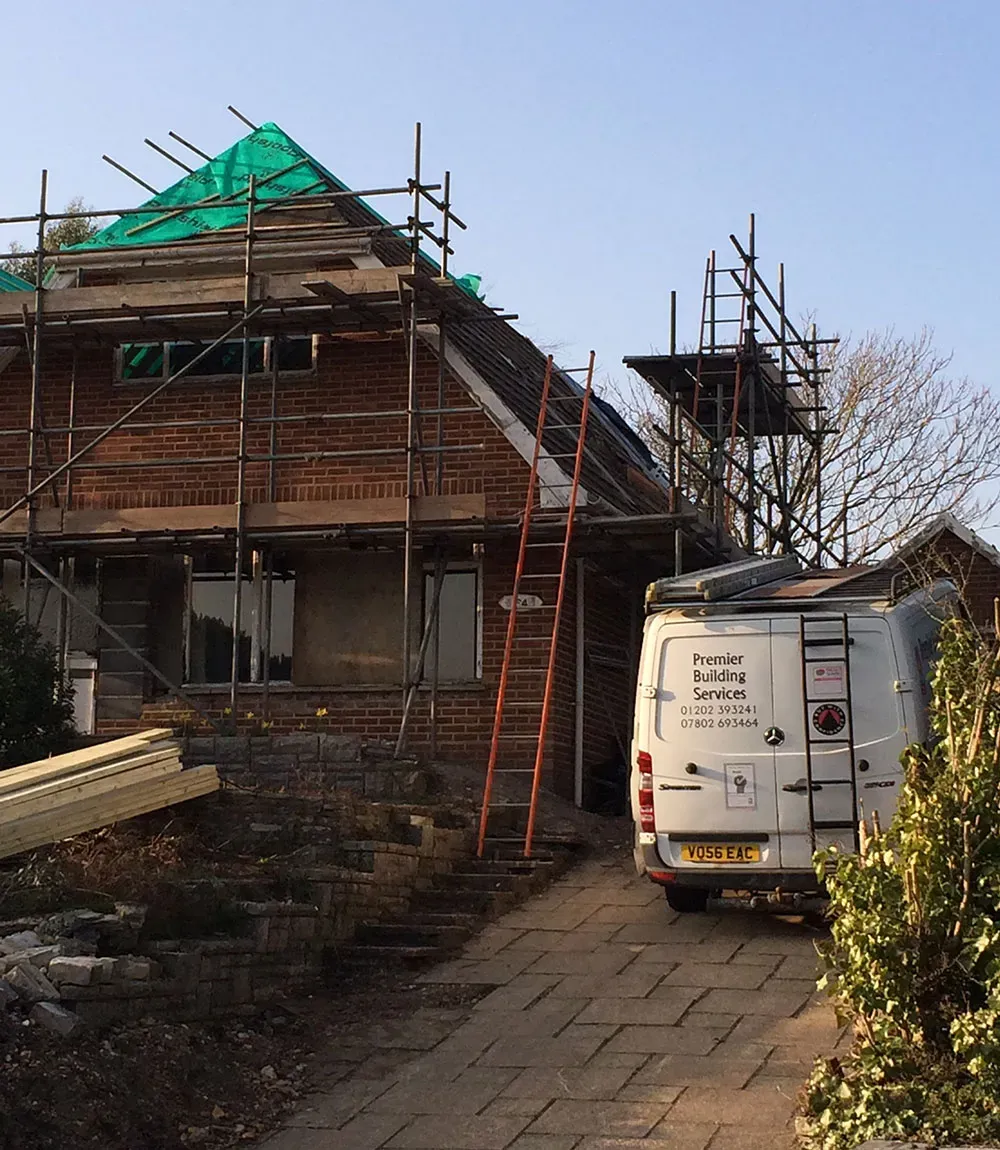 Brick house under scaffolding with its new roof wrapped in green membrane, and the Premier Building Services van parked on the drive