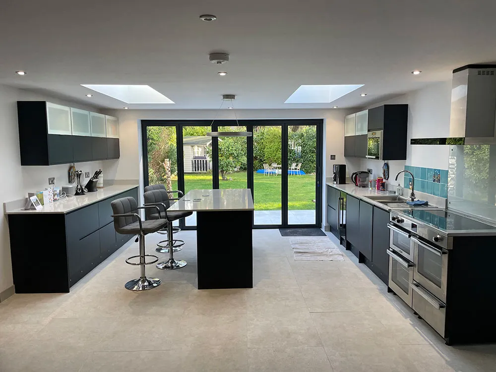 Kitchen after renovation: black fitted units, an island with bar stools and roof lanterns, looking out through bi-fold doors