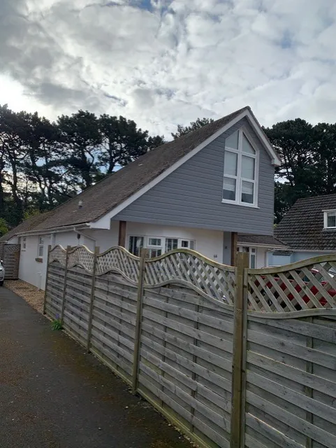 The finished grey clad gable end and side roof, seen from the lane beside the house over a timber fence