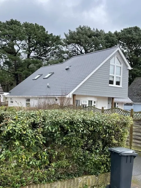 Side of the finished house seen over a hedge, with a grey clad gable, tall windows and roof windows in the grey tiled roof
